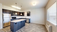 This well-appointed 2-bedroom, 2-bathroom residence at 2300 Main St in Washougal, WA offers a thoughtfully designed single-floor layout with a comfortable sense of space throughout. The open-concept kitchen and living area serves as the heart of the home, featuring striking navy cabinetry with brushed nickel hardware, light quartz countertops, a central island with pendant lighting, and a full suite of stainless steel appliances including a refrigerator, range, microwave, and dishwasher. Wood-look flooring runs throughout the kitchen and living areas, lending warmth and continuity. The spacious living room is bathed in natural light from a large sliding glass door that opens to a private balcony. Both bedrooms are carpeted and equipped with ceiling fans and windows that invite natural light. The generously sized primary bedroom includes a walk-in closet with wire shelving for ample storage. The two bathrooms are finished with wood-look tile flooring and dark cabinetry; one features a tub-shower combination and the other a walk-in shower. A hallway connects the private spaces, completing this clean, modern, and functional layout.