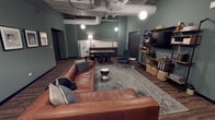 This single-floor space showcases a thoughtfully designed open-concept layout that blends industrial and modern aesthetics seamlessly. The living area anchors the space with a generous brown leather sectional sofa, complemented by a patterned area rug and a sleek cylindrical coffee table. A cozy accent chair in deep teal adds a pop of color alongside a minimalist side table, creating an inviting conversation area. Exposed HVAC ductwork runs across the ceiling, lending an industrial character that is balanced by warm pendant globe lights and a standing floor lamp. The walls are painted in a muted sage green, contributing to the calm, contemporary ambiance throughout. Toward the rear, a compact kitchenette or wet bar area features a countertop, sink, and seating for casual dining or meetings. A wall-mounted flat-screen TV is positioned on a dark open-shelving unit stocked with books, décor, and storage baskets, adding both function and visual interest. Dark hardwood-style flooring runs throughout, grounding the space with warmth and continuity. The overall design reflects a versatile, well-curated environment suited for lounging, socializing, or collaborative gatherings.