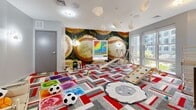 Located at 1212 Main St in Hartford, CT, this dedicated children's playroom is a thoughtfully designed, single-level space built to inspire creativity and active play. The room's most striking feature is a bold, floor-to-ceiling baseball-themed mural that dominates one wall, creating an immersive and energetic atmosphere. Complementing this are whimsical cloud-shaped wall sconces and pendant lights suspended from the ceiling, casting a soft, playful glow throughout the room. The floor is covered in a vibrant geometric carpet in red, grey, and white, adding both visual interest and a safe, cushioned surface for play. A wooden indoor climbing structure with a built-in slide serves as the centerpiece for active play, accompanied by a rock-climbing balance board. An interactive peg board display mounted on the mural wall offers a creative, hands-on activity station. A small table and chairs area provides a dedicated space for arts and crafts or quiet activities. Large windows along one wall invite an abundance of natural light and offer views of the surrounding neighborhood. The room is generously proportioned, providing ample open floor space for children to move freely, making it a well-rounded and engaging environment for young residents.