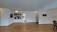 This single-floor space offers a clean, functional layout designed for comfortable everyday living. The main living area is characterized by smooth hardwood flooring that extends throughout, contributing warmth and continuity to the space. White walls and a standard ceiling with a ceiling fan and light fixture keep the area bright and well-ventilated.

A built-in white display cabinet with glass-paneled upper doors and solid lower cabinet doors serves as a notable architectural feature along one wall, providing both storage and display functionality. The cabinet is accented with decorative items, adding a touch of character to the otherwise neutral interior.

Two doorways lead from the main living area — one opening to what appears to be a kitchen, featuring white cabinetry and light countertops, and another leading to an entryway with a front door. The transitions between rooms are clearly defined, offering a straightforward and accessible floor plan.

The overall ambiance of the space is understated and practical, with a neutral color palette of whites and light wood tones that creates a versatile backdrop suitable for a variety of interior styles. The open feel of the main living area, combined with the natural flow between rooms, makes this a well-organized and approachable single-floor layout.
