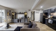 This well-appointed single-floor residence blends contemporary style with functional design throughout its thoughtfully arranged layout. The open-concept living area serves as the heart of the home, featuring light wood-look flooring, a textured ceiling, and a sliding glass door that opens to a private balcony, flooding the space with natural light. A rustic-modern entertainment center anchors the living room, complemented by a plush sectional seating arrangement and a marble-top coffee table set over a dark shag area rug.

The kitchen flows seamlessly from the living area and showcases bold dark cabinetry with gold hardware, black appliances, a white subway tile backsplash, and light countertops — creating a striking contrast that gives the space a modern edge.

The primary bedroom offers plush carpeting, a large sliding window with outdoor views, wall-mounted shelving, and a walk-in closet with built-in shelving and hanging rods. A second bedroom provides additional carpeted comfort with ample wall space and a dedicated closet. The bathroom features a floating dark vanity with a vessel sink, a large round mirror, a built-in vanity desk area, and a combined tub and shower, all finished with wood-look flooring for a cohesive aesthetic.