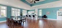 This single-floor clubhouse is defined by its cohesive design, featuring rich hardwood flooring and distinctive teal walls accented by crisp white crown molding and baseboards throughout. Natural light flows generously through numerous white-framed windows and sets of French doors that open to outdoor views, creating a bright and welcoming atmosphere across the entire space.

The lounge area serves as a central gathering point, anchored by a stone-surround fireplace topped with a white mantel and wall-mounted television. Dark leather sofas and armchairs furnish the area comfortably, while the French doors nearby provide easy outdoor access. Recessed lighting and additional wall sconces ensure the space remains well-lit at all times.

Adjacent to the lounge, the open dining and kitchen area features a tray ceiling with a ceiling fan, adding architectural depth. A large dark wood dining table with seating for multiple guests occupies the dining zone, while the kitchen includes white cabinetry, a kitchen island with bar-height seating, stainless steel appliances, and pendant lighting. A separate flex area with high-top bar seating and additional French doors rounds out this versatile, multi-functional space.
