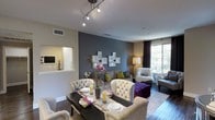 This single-floor space showcases a thoughtfully designed open-concept layout where the dining and living areas flow seamlessly together. The living and dining zone is anchored by rich hardwood-style flooring and a neutral color palette, punctuated by a striking slate-blue accent wall that adds depth and visual interest. A set of decorative mirror-framed wall panels serves as a focal point above the seating area, complementing the contemporary aesthetic.

The dining area features a sleek dark dining table surrounded by tufted, upholstered chairs with nailhead trim detailing, lending a refined, classic touch. A decorative table runner and floral centerpiece complete the setting. Adjacent to the dining space, the living area is furnished with coordinating tufted seating and a bold purple ottoman, providing vibrant contrast against the neutral tones.

Overhead, a track lighting fixture provides focused illumination throughout the combined space. Natural light filters in through a large window dressed with floor-length draping curtains, offering outdoor views. A convenient pass-through window connects to the kitchen area, which is visible in the background and features white cabinetry. The overall ambiance is modern, curated, and welcoming.