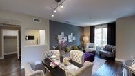This single-floor space showcases a thoughtfully designed open-concept layout where the dining and living areas flow seamlessly together. The living and dining zone is anchored by rich hardwood-style flooring and a neutral color palette, punctuated by a striking slate-blue accent wall that adds depth and visual interest. A set of decorative mirror-framed wall panels serves as a focal point above the seating area, complementing the contemporary aesthetic.

The dining area features a sleek dark dining table surrounded by tufted, upholstered chairs with nailhead trim detailing, lending a refined, classic touch. A decorative table runner and floral centerpiece complete the setting. Adjacent to the dining space, the living area is furnished with coordinating tufted seating and a bold purple ottoman, providing vibrant contrast against the neutral tones.

Overhead, a track lighting fixture provides focused illumination throughout the combined space. Natural light filters in through a large window dressed with floor-length draping curtains, offering outdoor views. A convenient pass-through window connects to the kitchen area, which is visible in the background and features white cabinetry. The overall ambiance is modern, curated, and welcoming.