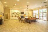 Located at 14332 Deloice Crescent in Newport News, Virginia, this single-floor space offers a refined and welcoming atmosphere characterized by thoughtful architectural details and generous, open layouts. Throughout the interior, large-format travertine tile flooring creates a cohesive, upscale aesthetic that flows seamlessly from room to room.

The main gathering area is a spacious, light-filled room anchored by a classic fireplace flanked by built-in white shelving with display lighting. A chandelier overhead adds warmth and elegance, while crown molding and recessed lighting contribute to the polished finish. An adjacent lounge area features a bay window arrangement surrounded by upholstered armchairs and a central coffee table beneath an orb chandelier, creating an intimate conversational setting bathed in natural light.

A large secondary common room opens generously to the outdoors through tall French doors, allowing ample natural light to fill the space. This room accommodates multiple seating arrangements, including tufted sofas, accent chairs, and dining-style chairs, all set atop area rugs that define each zone. Arched entryways and columned transitions add architectural character throughout.

A service counter area features white cabinetry with granite countertops, offering a practical refreshment station. The overall ambiance of the space is elegant yet approachable, blending classical design elements with comfortable, functional furnishings.