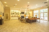 Located at 14332 Deloice Crescent in Newport News, Virginia, this single-floor space offers a refined and welcoming atmosphere characterized by thoughtful architectural details and generous, open layouts. Throughout the interior, large-format travertine tile flooring creates a cohesive, upscale aesthetic that flows seamlessly from room to room.

The main gathering area is a spacious, light-filled room anchored by a classic fireplace flanked by built-in white shelving with display lighting. A chandelier overhead adds warmth and elegance, while crown molding and recessed lighting contribute to the polished finish. An adjacent lounge area features a bay window arrangement surrounded by upholstered armchairs and a central coffee table beneath an orb chandelier, creating an intimate conversational setting bathed in natural light.

A large secondary common room opens generously to the outdoors through tall French doors, allowing ample natural light to fill the space. This room accommodates multiple seating arrangements, including tufted sofas, accent chairs, and dining-style chairs, all set atop area rugs that define each zone. Arched entryways and columned transitions add architectural character throughout.

A service counter area features white cabinetry with granite countertops, offering a practical refreshment station. The overall ambiance of the space is elegant yet approachable, blending classical design elements with comfortable, functional furnishings.