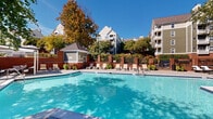 This outdoor amenity area is centered around a generously sized, crystal-clear swimming pool with vibrant turquoise water, designed to accommodate both relaxing swims and leisurely lounging. The pool deck is finished in a light-toned concrete surface and is lined with a collection of sling-style lounge chairs and chaise lounges, providing ample space for sunbathing and outdoor relaxation.

A charming gazebo-style poolhouse structure anchors one end of the deck, offering a shaded retreat with a pitched roof and classic architectural detailing. On the opposite side, a white pergola provides additional covered seating beneath shade trees, furnished with patio tables and chairs suitable for outdoor dining or socializing. Decorative brick columns and wrought iron fencing define the perimeter of the space, lending a traditional and refined aesthetic while maintaining an open, airy feel.

Mature trees throughout the area cast dappled natural light across the deck, creating a comfortable and inviting atmosphere during warm seasons. Vibrant flowering plants and lush greenery are strategically placed around the brick columns and fence line, enhancing the visual appeal of the space. The surrounding multi-story residential buildings provide an enclosed, community-oriented setting. Overall, the space is thoughtfully designed to offer both active and leisurely outdoor enjoyment.