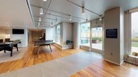 The game room amenity space at 770 Jackson Ave in Hoboken, New Jersey is a thoughtfully designed, single-level recreational facility offering a diverse range of entertainment and social areas. The layout flows seamlessly across warm hardwood flooring throughout, uniting each distinct zone into a cohesive, inviting environment.

The media lounge serves as a standout feature, appointed with a large-format flat-screen television mounted against a striking watercolor-style accent wall in soft teal tones. Comfortable sectional seating and a coffee table create an ideal setting for relaxed gatherings. Adjacent to this area, a dedicated games zone houses a full-size foosball table and a ping-pong table, complemented by lounge seating and curated wall art that lend the space a polished yet playful character.

The kitchenette is well-equipped with stainless steel appliances, including a full-size refrigerator, a sink with marble-style countertops, and a combination of upper white cabinetry and lower wood-toned cabinets — offering convenient refreshment options for users of the space.

Track lighting and recessed fixtures illuminate the games area, while large floor-to-ceiling windows flood the space with natural light and provide direct access to an outdoor terrace, extending the usable area and connecting the interior to the open air. The overall ambiance is modern, clean, and energetic — purpose-built for community recreation.