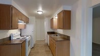 This single-floor space is thoughtfully laid out to maximize functionality and comfort. The kitchen serves as a central highlight, designed in a classic galley configuration that makes efficient use of the available space. Warm honey-toned wood cabinetry lines both sides of the kitchen, offering generous upper and lower storage for all culinary essentials. The dark laminate countertops provide a grounding contrast to the lighter cabinetry, while a double-basin stainless steel sink sits beneath under-cabinet lighting for added convenience. White appliances, including a freestanding gas range and a full-size refrigerator, complete the kitchen's practical setup. The floor is finished in neutral earth-toned ceramic tiles, adding durability and easy maintenance underfoot. Flush-mounted ceiling lighting provides consistent illumination throughout. A hallway extending from the kitchen leads further into the residence, hinting at additional living areas beyond. A window at the end of the hall allows natural light to filter into the space, brightening the interior. Overall, the space presents a clean, unpretentious aesthetic with a focus on practicality and efficient use of every square foot.