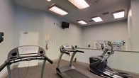 This single-floor fitness room offers a functional and well-equipped space designed for cardio-focused workouts. The room features light blue walls that contribute to a clean, open atmosphere, complemented by a gray carpeted floor that provides cushioning underfoot. Overhead, multiple recessed fluorescent light panels ensure consistent, bright illumination throughout the space.

The cardio equipment lineup includes at least two treadmills and an elliptical trainer, providing users with varied low-impact and moderate-intensity workout options. A wall-mounted flat-screen television is positioned for easy viewing during workouts, supported by an adjustable arm bracket. A full-length mirror along one wall visually expands the space and allows users to monitor their form during exercise.

Practical amenities include a fire extinguisher mounted on the wall and a thermostat for climate control, ensuring both safety and comfort. Ventilation is supported by ceiling vents integrated into the layout. The overall design is straightforward and utilitarian, prioritizing functionality and ease of use, making it well-suited as a shared or community fitness facility.