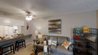 This single-floor space offers a welcoming and well-appointed interior designed for comfortable everyday living. The living area serves as the heart of the home, featuring plush carpeted flooring and a textured ceiling equipped with a ceiling fan with integrated lighting, ensuring year-round comfort. The room is furnished with a classic gray sofa and matching armchair, accented by colorful decorative pillows that add a vibrant, eclectic touch to the space. A small round side table with a table lamp provides warm ambient lighting, complementing the soft, neutral wall tones throughout. A bold, colorful abstract canvas artwork adorns one wall, serving as a striking focal point and adding personality to the room. A built-in or attached bar-height counter along one wall hints at a convenient pass-through or breakfast bar area, adding functionality to the layout. A metal shelving unit along the opposite wall offers additional display and storage space. The overall ambiance is warm, inviting, and casually styled, with thoughtful décor choices that balance comfort and character throughout the single-level floor plan.