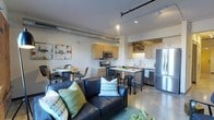 This single-floor space showcases a thoughtfully designed open-concept layout that blends living, dining, and kitchen areas into one cohesive environment. The industrial-chic aesthetic is immediately apparent through the exposed cylindrical ductwork running along the ceiling, complemented by a track lighting system that provides flexible, adjustable illumination throughout the space.

The living area anchors the space with a dark leather sofa accented by colorful throw pillows, creating a comfortable and stylish focal point. Adjacent to it, a compact dining nook accommodates a round table with modern chairs, making efficient use of the available floor space.

The kitchen is well-appointed with light wood cabinetry, a subway tile backsplash, and stainless steel appliances, including a full-size refrigerator, microwave, and range. A peninsula with bar seating provides additional prep space and casual dining options. A wine rack built into the upper cabinetry adds a distinctive functional detail.

Polished concrete floors flow continuously throughout, reinforcing the modern industrial character of the space. The neutral white walls and ceiling keep the overall atmosphere bright and open, while the thoughtful mix of textures and materials adds visual warmth and depth.
