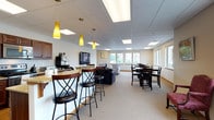 This single-floor community space is thoughtfully designed to accommodate a variety of social and functional needs. The open-concept layout flows seamlessly between distinct zones, each serving a specific purpose while maintaining a cohesive, welcoming atmosphere throughout.

The lounge area serves as a central hub, featuring a wall-mounted flat-screen television, bold patterned armchairs, and a tiled corner fireplace that adds warmth and comfort. Pendant lighting in a warm yellow hue accents the bar-height seating area, which is positioned near a coffee station and counter with a stone-topped surface, ideal for casual gatherings.

Adjacent to the lounge, a dedicated dining area offers ample seating around dark wood tables and chairs, bathed in natural light from large windows that overlook the surrounding neighborhood and parking area. The views provide a bright, airy feel to the space.

The fully equipped kitchen is a standout feature, outfitted with rich dark wood cabinetry, granite countertops, stainless steel appliances including a refrigerator, electric range, microwave, and dishwasher, as well as light hardwood-style flooring. This kitchen is well-suited for event preparation or community functions.

Throughout the space, neutral carpet and drop-panel ceilings with recessed lighting create a clean, comfortable environment suitable for a wide range of community activities.