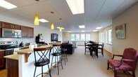 This single-floor community space is thoughtfully designed to accommodate a variety of social and functional needs. The open-concept layout flows seamlessly between distinct zones, each serving a specific purpose while maintaining a cohesive, welcoming atmosphere throughout.

The lounge area serves as a central hub, featuring a wall-mounted flat-screen television, bold patterned armchairs, and a tiled corner fireplace that adds warmth and comfort. Pendant lighting in a warm yellow hue accents the bar-height seating area, which is positioned near a coffee station and counter with a stone-topped surface, ideal for casual gatherings.

Adjacent to the lounge, a dedicated dining area offers ample seating around dark wood tables and chairs, bathed in natural light from large windows that overlook the surrounding neighborhood and parking area. The views provide a bright, airy feel to the space.

The fully equipped kitchen is a standout feature, outfitted with rich dark wood cabinetry, granite countertops, stainless steel appliances including a refrigerator, electric range, microwave, and dishwasher, as well as light hardwood-style flooring. This kitchen is well-suited for event preparation or community functions.

Throughout the space, neutral carpet and drop-panel ceilings with recessed lighting create a clean, comfortable environment suitable for a wide range of community activities.