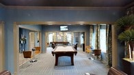 This single-floor amenity space is thoughtfully designed as a social and recreational hub. The room is anchored by a full-size billiards table positioned at the center, providing a focal point for leisure and entertainment. The interior is painted in a rich, muted blue palette that lends the space a calm yet sophisticated atmosphere, complemented by gold-toned trim accents framing the architectural columns and ceiling borders.

A wall-mounted flat-screen television is positioned for easy viewing from multiple areas of the room. Seating is distributed throughout in the form of sleek bistro-style tables paired with modern side chairs, ideal for casual conversation or relaxation. Decorative wall art, including circular metallic pieces, adds visual interest to the walls.

Large windows dressed with patterned curtains in earthy tones allow natural light to filter into the space, enhancing the open and airy feel. A striped, multi-toned carpet adds texture and definition to the floor. Additional recreational elements, including what appears to be a dartboard wall and a foosball or game table in the background, suggest this space is comprehensively equipped for a variety of social activities. Overall, the design strikes a balance between comfort, style, and functionality.