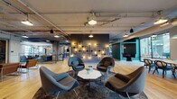 Located at 1300 N Fordham Blvd in Chapel Hill, NC, this single-floor amenity space combines industrial architecture with curated, eclectic interior design to create a dynamic and inviting communal environment. Exposed ductwork and concrete structural elements are balanced by warm hardwood flooring and carefully selected furnishings throughout.

The main lounge area features a striking dark accent wall adorned with an artistic pipe-and-bulb light installation, complemented by iconic womb-style chairs arranged around a marble-top coffee table. Large floor-to-ceiling windows flood the space with natural light and offer outdoor views, while a long communal dining table with pendant lighting provides a dedicated gathering spot.

A secondary lounge showcases rich tufted sofas, statement antler chandeliers, and bold patterned carpeting, lending a more intimate, layered ambiance. Adjacent to this is a well-appointed kitchen and social bar area featuring a polished concrete floor, shiplap wood ceiling, decorative tile accents, pendant lighting, built-in shelving, and a large central island with stylish bar seating.

Additional areas include a library nook with a floor-to-ceiling bookshelf, glass-railed mezzanine transition, and textured wallcoverings throughout. The space offers a cohesive blend of industrial, modern, and eclectic design elements suited for both relaxation and social interaction.