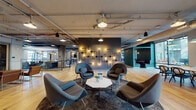 Located at 1300 N Fordham Blvd in Chapel Hill, NC, this single-floor amenity space combines industrial architecture with curated, eclectic interior design to create a dynamic and inviting communal environment. Exposed ductwork and concrete structural elements are balanced by warm hardwood flooring and carefully selected furnishings throughout.

The main lounge area features a striking dark accent wall adorned with an artistic pipe-and-bulb light installation, complemented by iconic womb-style chairs arranged around a marble-top coffee table. Large floor-to-ceiling windows flood the space with natural light and offer outdoor views, while a long communal dining table with pendant lighting provides a dedicated gathering spot.

A secondary lounge showcases rich tufted sofas, statement antler chandeliers, and bold patterned carpeting, lending a more intimate, layered ambiance. Adjacent to this is a well-appointed kitchen and social bar area featuring a polished concrete floor, shiplap wood ceiling, decorative tile accents, pendant lighting, built-in shelving, and a large central island with stylish bar seating.

Additional areas include a library nook with a floor-to-ceiling bookshelf, glass-railed mezzanine transition, and textured wallcoverings throughout. The space offers a cohesive blend of industrial, modern, and eclectic design elements suited for both relaxation and social interaction.
