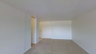 This single-floor residential space offers a clean and functional layout designed with simplicity and practicality in mind. The interior is characterized by crisp white walls that reflect light evenly throughout, contributing to a bright and open atmosphere. The flooring is covered with wall-to-wall carpet in a neutral tone, providing a soft underfoot feel that extends across the main living area. The ceiling features a classic textured finish, adding a subtle layer of visual detail overhead. The main living area is generously proportioned, offering ample space for a variety of furniture arrangements and uses. A distinct architectural nook or alcove is visible along one wall, providing a natural division within the open floor plan and offering potential for flexible use. The entryway, accessible through a solid door, is finished with tiled flooring, creating a practical transition zone between the exterior and the carpeted interior. The overall ambiance of the space is understated and move-in ready, presenting a neutral canvas well-suited for personalization. The straightforward design and consistent finishes throughout make this a highly adaptable space for a range of occupants and lifestyle needs.