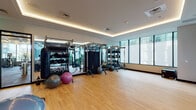 This contemporary fitness center is thoughtfully designed to support a wide range of training styles within a bright, open environment. The space features warm hardwood flooring throughout, providing a clean and durable surface ideal for both equipment-based and open-floor workouts. Recessed cove lighting along the tray ceiling casts a warm, ambient glow that complements the abundant natural light streaming in through floor-to-ceiling windows along the perimeter walls, offering views of the surrounding urban landscape.

The facility is outfitted with a comprehensive selection of functional training equipment, including a multi-station cable and functional training rig, TRX suspension trainers, a full rack of kettlebells and dumbbells, stability balls, medicine balls, plyo boxes, and a heavy punching bag. Full-length mirrors are positioned along one wall, enhancing the sense of depth and allowing users to monitor form during exercise. A wall-mounted display screen serves as a focal point for motivational content or guided workouts.

A standout architectural feature is the glass-walled connection to a landscaped outdoor terrace, accessible through double glass doors. The terrace is furnished with hanging swing chairs and lush planters, creating a tranquil retreat directly adjacent to the workout floor. This seamless indoor-outdoor integration adds a distinctive, resort-like quality to the overall fitness experience.