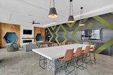 Located at 2225 Collett Ave in Corona, California, this single-floor amenity space offers a dynamic and thoughtfully designed lounge environment that seamlessly blends social and recreational functions. The space is anchored by a striking accent wall featuring a bold geometric pattern in charcoal and lime green, which immediately sets a vibrant, contemporary tone throughout.

The living area is furnished with a generous sectional sofa accented by colorful throw pillows, centered around a round coffee table and positioned in front of a sleek linear fireplace mounted on a warm wood-paneled wall. A unique hexagonal nook with tufted blue velvet seating adds a playful architectural detail, while a wall-mounted flat-screen TV completes the entertainment setup. A ceiling fan with a tray ceiling and ambient cove lighting adds comfort and polish to the room.

Large floor-to-ceiling sliding glass doors flood the living area with natural light and open to an outdoor area with pool views, creating a seamless indoor-outdoor connection. A foosball table positioned centrally adds a recreational element to the lounge.

The dining and kitchen area features a long white countertop bar with leather barstools, warm wood cabinetry, open floating shelves with curated décor, and pendant lighting overhead. Large-format hexagonal tile flooring unifies the entire space with a clean, modern aesthetic.