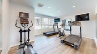 This single-floor amenity facility in Tulsa, Oklahoma offers two functional spaces designed to serve residents with convenience and comfort. The layout connects an exercise room and a laundry room via a central hallway, all finished with light-toned luxury vinyl plank flooring that gives the space a clean, contemporary feel throughout.

The exercise room is bright and inviting, benefiting from large windows that overlook an outdoor pool area, allowing natural light to fill the space. It is equipped with cardio machines including treadmills, an elliptical, and a stationary bike, each outfitted with individual monitors. Wall-mounted flat-screen TVs and mirrored wall panels enhance the workout environment, making the room feel open and well-appointed despite its compact footprint.

The laundry room is neatly organized with multiple washers and dryers lining the walls on both sides, providing ample capacity. Recessed lighting illuminates the space evenly, and light gray walls contribute to a tidy, neutral aesthetic. A wall-mounted cabinet, informational bulletin board, and centrally placed waste bin add practical touches to the room.

Both spaces feature recessed ceiling lighting and HVAC vents, ensuring a comfortable environment year-round. The overall design is functional, modern, and well-maintained, reflecting a thoughtfully managed shared amenity facility.