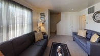 This two-story home offers a practical and comfortable layout spread across two levels, connected by a carpeted staircase visible from the living area. The main level features a living room with plush carpeting, natural light from a large window with sheer curtains, and a welcoming atmosphere. Adjacent to the living area, a dedicated dining space accommodates a round table for four and includes a ceiling fan with light fixture, a window, and direct access to an entry door.

The kitchen is arranged in a U-shaped configuration, providing ample counter space with light-toned laminate countertops, white cabinetry, a stainless steel sink beneath a window, and includes a dishwasher and range.

A bathroom on the upper level features a single vanity with mirror, and a tub-shower combination with a bold chevron-patterned curtain. An additional space includes grab bar hardware, indicating accessibility-conscious design.

The home includes multiple bedrooms, each carpeted and filled with natural light from windows dressed with curtains. The bedrooms offer generous floor space suitable for full or queen-sized beds along with storage furniture. The overall palette throughout is neutral, with warm cream-toned walls creating a consistent and cohesive ambiance across all rooms.