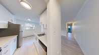 This single-floor unit presents a clean, contemporary design characterized by crisp white walls, light wood-toned flooring, and abundant natural light throughout. The kitchen is thoughtfully appointed with white shaker-style cabinetry extending to the ceiling, stone-look countertops, a stainless steel sink, a built-in dishwasher, and a full-size stainless steel refrigerator. White subway tile backsplash adds a polished finish, while a window above the counter brings in natural light and views of the surrounding brick residential buildings. A central hallway connects the kitchen and living areas to the private rooms, maintaining a logical and efficient flow. The living and bedroom spaces are generously proportioned, each featuring large sliding or casement windows fitted with baseboard heating units beneath — ensuring comfort throughout the seasons. Walls are painted in a neutral white palette that maximizes the sense of brightness and openness. The bathroom is finished with large-format white wall tiles, a soaking tub with an overhead shower, chrome fixtures, and a window for natural ventilation. Light wood flooring runs consistently throughout the unit, unifying the overall aesthetic. The space is move-in ready, offering a modern and well-maintained environment suited for comfortable everyday living.