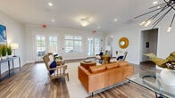 This well-appointed single-floor space combines a clean, coastal-inspired aesthetic with practical, welcoming design. The main lounge area serves as the heart of the space, centered around a rich cognac leather sofa paired with natural wood-framed armchairs upholstered in crisp white fabric and accented with navy blue throw pillows. A round woven gold sunburst mirror adds a decorative focal point to the soft gray walls, complemented by recessed lighting, a ceiling fan, and elegant crown molding throughout.

Wide-plank hardwood-style flooring extends seamlessly across the entire space, lending warmth and cohesion from room to room. Multiple sets of white-framed French doors line the walls, flooding the interior with natural light and offering views of the surrounding outdoor area. These doors provide easy indoor-outdoor connectivity and contribute to the open, airy atmosphere of the space.

A transitional entryway welcomes visitors with a sleek console table, framed artwork depicting vibrant blue skies, and decorative greenery. Hallways connect additional rooms, including what appears to be a private office or conference space with task chairs visible through an open doorway. The overall ambiance is bright, polished, and inviting, reflecting a carefully curated blend of modern comfort and casual elegance suited to a communal or professional setting.