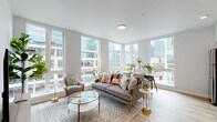 This single-floor urban residence offers a thoughtfully designed open-concept layout bathed in natural light. The living area is a standout, featuring expansive floor-to-ceiling windows on multiple walls that frame sweeping city skyline views. Light hardwood flooring runs throughout, complementing the neutral, airy palette. The living space comfortably accommodates a seating arrangement centered around a glass-top coffee table, with room for a dining area near the windows.

The kitchen is seamlessly integrated into the open living space and features sleek white cabinetry extending to the ceiling, light-toned tile backsplash, white quartz countertops, stainless steel appliances, and an undermount sink. Open shelving adds a contemporary touch and visual depth.

The bedroom offers a more intimate atmosphere, highlighted by a dramatic custom wall mural in deep purple and gold tones that serves as an artistic focal point behind the tufted upholstered headboard. A large window allows natural light and city views into the room. Brass-accented nightstands add warmth and sophistication.

The bathroom maintains the clean, modern aesthetic with a white vanity, quartz countertop, undermount sink, and a backlit LED mirror, all set against soft neutral walls and hardwood-style flooring.