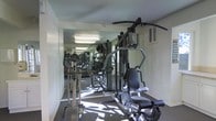 This single-floor fitness center is designed to provide a functional and efficient workout environment. The space is outfitted with a selection of exercise equipment, including a multi-station cable and weight machine, an elliptical trainer, and additional cardio and strength machines that cater to a range of fitness needs. A large wall-to-wall mirror runs along one side of the room, visually expanding the space and allowing users to monitor their form during exercise. The reflective surface also amplifies the natural light that filters into the room, contributing to a bright and energizing atmosphere. The flooring is finished with a durable, dark-toned carpet well-suited to a gym environment. Overhead fluorescent lighting ensures consistent illumination throughout the space. A ventilation unit is mounted on the upper wall, supporting adequate airflow during workouts. The layout is thoughtfully arranged to maximize usable floor space, allowing for comfortable movement between stations. Overall, this fitness center offers a practical and straightforward setting for residents or guests looking to maintain an active lifestyle without leaving the building.
