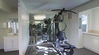 This single-floor fitness center is designed to provide a functional and efficient workout environment. The space is outfitted with a selection of exercise equipment, including a multi-station cable and weight machine, an elliptical trainer, and additional cardio and strength machines that cater to a range of fitness needs. A large wall-to-wall mirror runs along one side of the room, visually expanding the space and allowing users to monitor their form during exercise. The reflective surface also amplifies the natural light that filters into the room, contributing to a bright and energizing atmosphere. The flooring is finished with a durable, dark-toned carpet well-suited to a gym environment. Overhead fluorescent lighting ensures consistent illumination throughout the space. A ventilation unit is mounted on the upper wall, supporting adequate airflow during workouts. The layout is thoughtfully arranged to maximize usable floor space, allowing for comfortable movement between stations. Overall, this fitness center offers a practical and straightforward setting for residents or guests looking to maintain an active lifestyle without leaving the building.