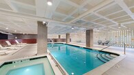 This contemporary indoor aquatic and wellness facility is thoughtfully designed to deliver a premium recreational experience. At its heart is a generously sized indoor lap pool with crystal-clear turquoise water, flanked by a hot tub and ample pool deck space. The pool area is defined by striking architectural columns, a coffered white ceiling with integrated track lighting, and a dramatic perforated block wall punctuated by colorful accent tiles that diffuse light throughout the space, creating a bright and welcoming atmosphere.

A dedicated lounge area adjoins the pool, furnished with sleek white sun loungers and side tables set against rich wood-paneled wainscoting beneath a deep navy accent wall, lending the space a refined, resort-like ambiance. The locker room area provides modern amenities including secure electronic lockers, open shower stalls lined with textured blue tile, and private changing areas finished in large-format stone-look porcelain tile.

The facility's entrance corridor features an eye-catching lobby with large-format grey tile flooring, glass-partitioned hallways, and curated planter installations filled with ornamental grasses and river stones, contributing a biophilic design element. Throughout the space, the material palette of concrete, polished tile, and glass creates a cohesive, contemporary aesthetic suited to a high-end fitness or residential amenity environment.