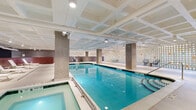 This contemporary indoor aquatic and wellness facility is thoughtfully designed to deliver a premium recreational experience. At its heart is a generously sized indoor lap pool with crystal-clear turquoise water, flanked by a hot tub and ample pool deck space. The pool area is defined by striking architectural columns, a coffered white ceiling with integrated track lighting, and a dramatic perforated block wall punctuated by colorful accent tiles that diffuse light throughout the space, creating a bright and welcoming atmosphere.

A dedicated lounge area adjoins the pool, furnished with sleek white sun loungers and side tables set against rich wood-paneled wainscoting beneath a deep navy accent wall, lending the space a refined, resort-like ambiance. The locker room area provides modern amenities including secure electronic lockers, open shower stalls lined with textured blue tile, and private changing areas finished in large-format stone-look porcelain tile.

The facility's entrance corridor features an eye-catching lobby with large-format grey tile flooring, glass-partitioned hallways, and curated planter installations filled with ornamental grasses and river stones, contributing a biophilic design element. Throughout the space, the material palette of concrete, polished tile, and glass creates a cohesive, contemporary aesthetic suited to a high-end fitness or residential amenity environment.