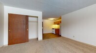 This single-floor unit offers a straightforward, functional layout connecting a carpeted living area to a compact kitchen. The living space features neutral beige carpeting, white walls, and a textured ceiling, creating a clean and simple atmosphere. A sliding-door closet provides convenient built-in storage directly off the main living area.

The kitchen transitions from the living space via tile flooring in a warm earth tone. Oak cabinetry lines the upper and lower walls, offering ample storage. White appliances, including a freestanding range, refrigerator, and a single-basin sink, complete the functional cooking area.

The bathroom is tiled in white from floor to wall, contributing a clean, utilitarian feel. It includes an oak vanity with an integrated sink, a wall-mounted mirror, and standard white fixtures. The tile flooring mirrors the earthy tones found in the kitchen.

The bedroom is carpeted and brightened by a window that allows natural light and views of surrounding trees. A baseboard heating system runs along the exterior wall, and the textured ceiling is consistent throughout the unit. Overall, the space is practical and move-in ready.