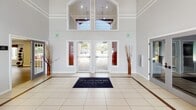 This single-story clubhouse, branded under McKinley, makes an immediate impression with its grand entrance foyer, where soaring double-height ceilings, arched transom windows, and large glass entry doors flood the space with natural light. Light tile flooring with contrasting border inlays runs throughout the main common areas, reinforcing the open, airy feel.

The central lounge area is a standout, featuring dramatic floor-to-ceiling windows that frame direct waterfront views, including an outdoor dock and open water beyond. Comfortable seating arrangements and a wall-mounted television create a welcoming social environment. An adjacent dining area with bistro-height seating offers additional gathering space with continued water views.

The fitness facilities are comprehensive, comprising a dedicated cardio and strength training room with rubberized flooring and modern equipment, as well as a separate open studio room with wood-look flooring, ceiling fans, and garden-facing windows stocked with medicine balls for group exercise or yoga.

A compact catering kitchen features dark wood cabinetry, granite countertops, stainless steel appliances, and wood parquet flooring. French doors connect the fitness areas to the main lounge, promoting seamless circulation throughout this well-appointed community amenity space.