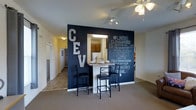 This single-floor unit makes a strong visual statement with a dramatic floor-to-ceiling chalkboard accent wall that serves as both a design focal point and a functional, writable surface. Mounted decorative block letters and handwritten chalk messaging add a personalized, community-oriented character to the space. A built-in pass-through window connects the living area to the kitchen, topped with a white breakfast bar that accommodates two bar-height chairs, making it ideal for casual dining or socializing. The kitchen, visible through the pass-through, features wood-toned cabinetry and includes a refrigerator, offering a fully equipped cooking space. The open layout flows seamlessly from the dining area into a carpeted living space, with a hallway extending toward additional rooms. A ceiling fan with a light fixture provides both air circulation and ambient lighting overhead, while track lighting near the accent wall adds focused illumination. The combination of dark navy walls against light carpet and white trim creates a bold contrast throughout the space, giving it an energetic and modern aesthetic well-suited to active, social living.