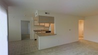 This single-floor space features an open-concept design that integrates the kitchen and living area into one cohesive layout. The kitchen is equipped with light wood cabinetry, a dark countertop peninsula that doubles as a breakfast bar, and a built-in range with a large overhead range hood as a focal point. A sink with a single faucet is positioned along the kitchen counter, and dark appliances are visible within the cooking area. Upper cabinets provide ample storage, and HVAC vents are positioned in the ceiling for efficient climate control throughout. The flooring transitions from tile in the kitchen zone to carpet in the living area, adding textural contrast. The living space is generously sized with neutral walls and a clean, minimalist aesthetic throughout. A hallway visible beyond the kitchen leads to additional rooms, suggesting a functional floor plan with clear separation between common and private areas. The overall ambiance is bright and understated, with neutral tones dominating the palette and practical finishes suited for everyday living.
