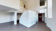 This two-story space makes a strong architectural impression through its dramatic double-height ceilings, which flood the interior with a sense of openness and volume. The main living area is anchored by dark hardwood flooring that runs throughout, providing a rich contrast against the crisp white walls and trim.

A central staircase, framed by clean white railings, serves as both a functional and visual focal point of the space. Cleverly integrated built-in shelving units are incorporated into the staircase structure, offering generous storage and display options while maintaining the streamlined aesthetic of the design.

An open upper-level mezzanine overlooks the main floor below, connected by the staircase and reinforcing the spacious, loft-like quality of the layout. Recessed HVAC vents are positioned throughout, ensuring practical climate control without disrupting the clean lines of the interior.

Toward the rear of the main floor, rich dark cabinetry hints at a kitchen or wet bar area, providing a warm accent against the predominantly neutral palette. Overall, the space combines contemporary design with thoughtful built-in features, creating a functional and visually dynamic environment.