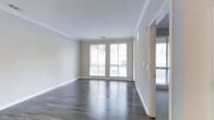 This single-floor space offers a clean, modern aesthetic defined by thoughtful finishes and an open, airy feel. The main living area showcases rich, dark hardwood flooring that runs throughout, providing a striking contrast against the neutral, warm-toned walls and crisp white trim. Elegant crown molding frames the ceilings, adding a refined architectural detail that elevates the overall design. Multiple large windows fitted with white blinds allow an abundance of natural light to pour into the space, creating a bright and welcoming atmosphere. The windows also offer views of lush greenery outside, bringing a sense of the outdoors in. Recessed lighting in the ceiling supplements the natural light, ensuring the space remains well-lit at all times. The layout feels open and spacious, with smooth, clean wall surfaces and generous proportions that offer flexibility for a variety of uses. Wide baseboards complement the crown molding, contributing to a cohesive and polished look throughout. The overall ambiance is one of understated elegance — move-in ready with a neutral palette that serves as an ideal backdrop for any interior style.