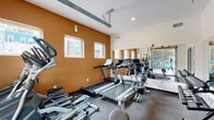 Located at 916 N College Ave in Bloomington, Indiana, this fitness center is a well-appointed exercise room designed to accommodate a full range of workout routines within a compact footprint. The space features a bold warm-orange accent wall that energizes the room, complemented by crisp white ceilings and walls that keep the atmosphere bright and motivating.

The cardio equipment lineup includes multiple treadmills and elliptical machines, providing ample options for aerobic training. A dedicated free weights area features a tiered dumbbell rack stocked with a complete set of weights, ideal for strength training. A full-wall mirror spans one side of the room, visually expanding the space and allowing users to monitor their form during workouts.

A standout architectural element is the large glass garage-style door, which opens to an outdoor area adorned with vibrant mural artwork, offering a distinctive visual backdrop and the option for fresh air. Multiple windows along the accent wall allow natural light to filter in and provide views of the surrounding neighborhood. The durable rubber flooring throughout is designed to withstand heavy equipment and high-traffic use. A coat hook wall panel near the entrance adds practical convenience. The overall design balances functionality and aesthetic appeal, creating an inviting environment for daily fitness activity.