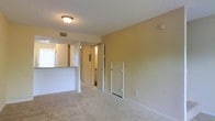 This two-floor residential space offers a practical and straightforward layout designed for comfortable everyday living. The main living area is finished with wall-to-wall carpet in a neutral beige tone, complementing the warm, light-colored walls throughout. A textured ceiling adds subtle dimension to the space, while a flush-mount ceiling light fixture provides overhead illumination.

A half-wall partition separates the kitchen area from the living space, creating a sense of openness while maintaining a defined boundary between the two zones. The kitchen area, visible beyond the divider, benefits from natural light filtering in through a window.

Along the far wall of the living area, multiple interior doors are visible — one leading to what appears to be a bathroom, identifiable by the lighting and fixtures glimpsed through the open door, and a smaller door likely serving as a utility or storage closet. A hallway connects these areas, providing clear circulation through the space.

Practical details such as HVAC vents, a smoke detector, and standard electrical outlets are integrated throughout, reflecting a functional residential design. The two-floor configuration further expands the overall living footprint of this space.