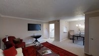 This single-floor residence offers a thoughtfully arranged layout that balances comfort and functionality. The living area serves as the heart of the space, featuring neutral-toned walls, plush carpeting, and a textured ceiling with classic crown molding details. A wall-mounted flat-screen television anchors the main wall, complemented by tasteful furnishings including a glass-top coffee table and a rich, red area rug that adds a warm pop of color to the room. Natural light filters softly through the space, contributing to an inviting ambiance. Adjacent to the living area, a defined dining nook provides an intimate setting for meals, furnished with a compact table and seating. A pass-through or open counter connects this area to the kitchen, promoting a seamless flow between spaces. The kitchen is visible through a doorway and features white cabinetry and appliances, offering a clean and efficient workspace. The overall design maintains a cohesive, neutral color palette throughout, creating a calm and welcoming atmosphere. The open sightlines between the living, dining, and kitchen areas make the space feel connected and easy to navigate, ideal for both relaxed daily living and casual entertaining.