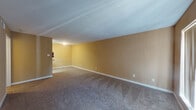 This single-floor unit offers a functional and straightforward layout across several connected rooms. The living areas are finished with neutral, warm-toned walls in a soft golden beige, complemented by wall-to-wall carpet in a medium brown tone that runs throughout the main rooms. White baseboards provide clean contrast along the perimeter of each space.

The main living area is generously sized, featuring an entry door alongside a large window fitted with vertical blinds that allow natural light to filter in. A secondary room, suitable as a bedroom, includes a ceiling fan with integrated lighting and a recessed window nook that adds architectural interest while bringing in additional natural light.

The galley-style kitchen is efficiently arranged with white cabinetry lining both sides, granite-look laminate countertops, a tile backsplash in earthy tones, and white appliances including a range and refrigerator. Wood-look flooring runs through the kitchen, adding warmth. A double sink sits opposite the appliances, and a closet at the end of the kitchen provides additional storage.

The bathroom features wood-look flooring, a full-size soaking tub with a shower, and white square wall tiles accented by a decorative mosaic border in brown tones. A fire extinguisher is mounted in the hallway, and multiple electrical outlets are distributed throughout the unit.