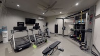 This two-floor fitness space is thoughtfully designed to accommodate a comprehensive range of workout disciplines. The lower level serves as the primary strength and cardio zone, featuring durable rubber flooring, recessed lighting, and a large ceiling fan paired with a wall-mounted mini-split system for climate control. Equipment includes multiple treadmills, an elliptical, a flat weight bench, a full dumbbell rack, and a wall-mounted flat-screen TV. An Octagon functional training rig anchors one wall, complete with a heavy punching bag, resistance attachments, and organized storage for medicine balls, battle ropes, and accessories. A dramatic spiral staircase with sleek black metal railings connects the two levels, serving as both a functional and architectural centerpiece.

The upper level transitions to lighter wood-look flooring and an airier atmosphere, also equipped with a ceiling fan and mini-split unit. This floor is dedicated to cycling and functional training, housing several Spinning bikes and balance and stability equipment. A large, vibrant wall mural featuring an abstract peacock design adds an energetic focal point, complemented by a wall-mounted TV and mirrored doors. Together, both floors create a cohesive, well-equipped private fitness environment.