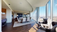 Located along Windrose Avenue in Plano, Texas, this high-rise residence offers a sophisticated single-floor layout defined by soaring ceilings and dramatic floor-to-ceiling curved glass walls that frame panoramic views of the North Dallas skyline and surrounding cityscape. Rich hardwood-style flooring flows throughout the main living areas, lending warmth to the contemporary design.

The open living and dining area is anchored by the sweeping curved glass facade, flooding the space with abundant natural light and providing a striking backdrop for everyday living and entertaining. A dedicated study or sitting room features open metal-and-wood shelving and direct access to the expansive city views.

The chef's kitchen is outfitted with dark wood cabinetry, stainless steel appliances, a professional-grade range, and a generously sized island topped with a distinctive veined stone countertop, complemented by large pendant lighting.

The primary bedroom suite enjoys its own wall of floor-to-ceiling windows and connects to a spa-inspired bathroom featuring a freestanding soaking tub positioned to capture the open skyline, dark wood vanities with dual sinks, and ample storage. Secondary living spaces offer carpeted comfort and continued city views. A dedicated laundry area with full-size washer and dryer adds practical convenience to this elegantly appointed high-rise residence.