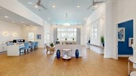 Located at 9505 Blackwood Circle in Fort Myers, FL, this single-floor community space offers an airy, coastal-inspired environment defined by soaring ceilings, crisp white walls, and warm wood-look flooring throughout. The open-concept layout seamlessly integrates the living and dining areas, creating a generous, welcoming atmosphere ideal for gathering and socializing.

The living area is anchored by a comfortable seating arrangement beneath a soft blue tray ceiling accented with recessed lighting and ceiling fans, which enhance both the aesthetic and comfort of the space. Large multi-pane windows flood the interior with natural light and offer views of the outdoor surroundings, while patterned draperies in a navy and white motif add a refined coastal touch.

The dining area features high-top tables and blue upholstered chairs positioned beneath floor-to-ceiling windows that frame views of the outdoor pool area. A glass-panel door provides direct access to the exterior patio, further blurring the line between indoor and outdoor living.

A well-appointed foyer with a built-in ledge, decorative console table, and interior window details creates an elegant entry sequence. A secondary area, visible from the main hall, showcases additional cabinetry and a starburst light fixture. Throughout the space, navy blue decorative accents, potted greenery, and coastal artwork contribute to a cohesive and polished Florida aesthetic.
