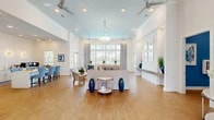 Located at 9505 Blackwood Circle in Fort Myers, FL, this single-floor community space offers an airy, coastal-inspired environment defined by soaring ceilings, crisp white walls, and warm wood-look flooring throughout. The open-concept layout seamlessly integrates the living and dining areas, creating a generous, welcoming atmosphere ideal for gathering and socializing.

The living area is anchored by a comfortable seating arrangement beneath a soft blue tray ceiling accented with recessed lighting and ceiling fans, which enhance both the aesthetic and comfort of the space. Large multi-pane windows flood the interior with natural light and offer views of the outdoor surroundings, while patterned draperies in a navy and white motif add a refined coastal touch.

The dining area features high-top tables and blue upholstered chairs positioned beneath floor-to-ceiling windows that frame views of the outdoor pool area. A glass-panel door provides direct access to the exterior patio, further blurring the line between indoor and outdoor living.

A well-appointed foyer with a built-in ledge, decorative console table, and interior window details creates an elegant entry sequence. A secondary area, visible from the main hall, showcases additional cabinetry and a starburst light fixture. Throughout the space, navy blue decorative accents, potted greenery, and coastal artwork contribute to a cohesive and polished Florida aesthetic.