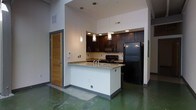 This single-floor space showcases a contemporary open-concept design with a cohesive industrial-modern aesthetic throughout. The kitchen serves as the focal point of the space, featuring rich dark wood cabinetry that contrasts elegantly against the crisp white walls and light granite countertops. A peninsula-style kitchen island provides additional workspace and casual seating, making it ideal for both meal preparation and entertaining. Black appliances, including a full-size refrigerator, range, and microwave, lend a sleek and uniform look to the culinary space. Stylish pendant lights hang above the island, adding a warm, focused glow that complements the recessed lighting overhead. The soaring ceiling height amplifies the sense of openness, creating an airy and expansive atmosphere. Polished concrete floors in a distinctive green-tinted finish run throughout, adding a bold industrial character that sets the space apart. A louvered wood door, likely leading to a utility or storage area, adds a warm natural accent near the kitchen entry. The overall ambiance balances modern functionality with distinctive design details, resulting in a space that feels both practical and visually striking.