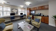 This business center offers a thoughtfully designed, single-floor space that seamlessly blends functionality with comfort. The layout is divided into two distinct zones: a dedicated computer workstation area and a relaxed lounge seating area, making it suitable for a variety of tasks.

The workstation zone features a long, sleek desk running along a wall of large windows, fitted with multiple desktop computers and blue accent chairs. Natural light filters through roller shades, creating a bright and focused work environment with views of the surrounding exterior.

The lounge area is furnished with plush, gold-toned wingback chairs accented with navy blue patterned throw pillows, arranged around a modern rectangular coffee table. Curated artwork, including a bold blue abstract piece, adds a sophisticated visual element to the neutral-toned walls. Decorative accents and coffee table books complete the refined aesthetic.

A built-in cabinetry unit with warm wood tones houses a flat-screen television and coffee station, offering added convenience. Drum-style flush-mount ceiling fixtures provide consistent, warm overhead lighting throughout. Glass-paneled French doors connect the business center to adjacent common areas, contributing to an open and accessible feel. The space is finished with neutral carpet and crisp white millwork, creating a professional yet inviting atmosphere.