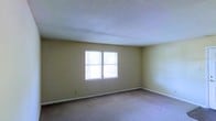 This single-floor space offers a clean and straightforward layout designed for comfortable, everyday living. The main room is finished with wall-to-wall carpet in a neutral tone, providing a soft underfoot feel throughout. The walls are painted in a warm, understated beige, creating a calm and inviting atmosphere that serves as a versatile backdrop for a variety of furnishing styles. A textured popcorn ceiling adds a classic touch overhead, while white baseboard trim runs along the perimeter of the room, offering a clean visual boundary between the walls and floor. A centrally positioned window allows natural light to filter into the space, brightening the interior and offering a view of the surrounding greenery outside. The window's placement ensures the room feels open and airy despite its simple design. Overall, the space presents a modest yet functional environment, well-suited for those seeking a straightforward, move-in-ready layout with a neutral palette that can easily be personalized to suit individual tastes.
